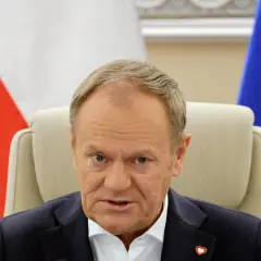 Donald Tusk, z flagą Polski i UE za plecami, w białej koszuli i granatowej marynarce. Czy Donald Tusk ma pochodzenie niemieckie?