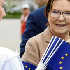 Ewa Kopacz, gdzie mieszka, uśmiecha się, trzymając flagi UE.