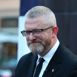Grzegorz Braun, w okularach i z brodą, w czarnym garniturze, z mikrofonem przypiętym do klapy.