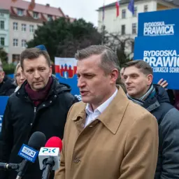 Mężczyzna w płaszczu przemawia do mikrofonów. W tle ludzie z banerami "Bydgoska Prawica". Chcesz wiedzieć, jak zostać politykiem Konfederacji?