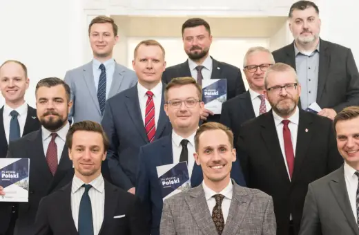 Grupa mężczyzn w garniturach, jeden trzyma broszurę "Po stronie Polski". To członkowie Konfederacji, partii politycznej.