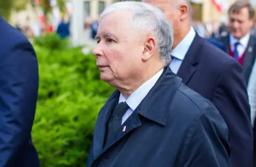 Jarosław Kaczyński, lider partii, w czarnym płaszczu i z przypinką z polską flagą, podczas oficjalnych uroczystości.
