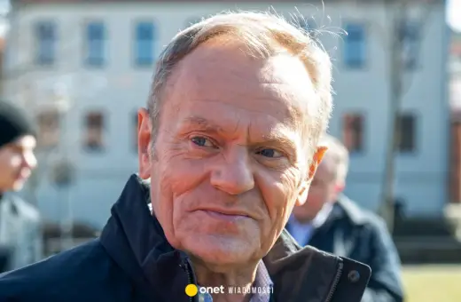 Zbliżenie na twarz Donalda Tuska. W tle widać niewyraźne sylwetki innych osób i budynek. Wiele osób zastanawia się, ile lat ma Tusk, a jego wygląd sugeruje doświadczenie.