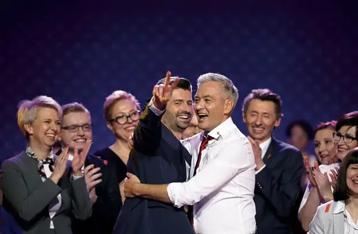 Dwóch mężczyzn, jeden z nich to partner Biedronia, ściska się na scenie, podczas gdy inni ludzie klaszczą.