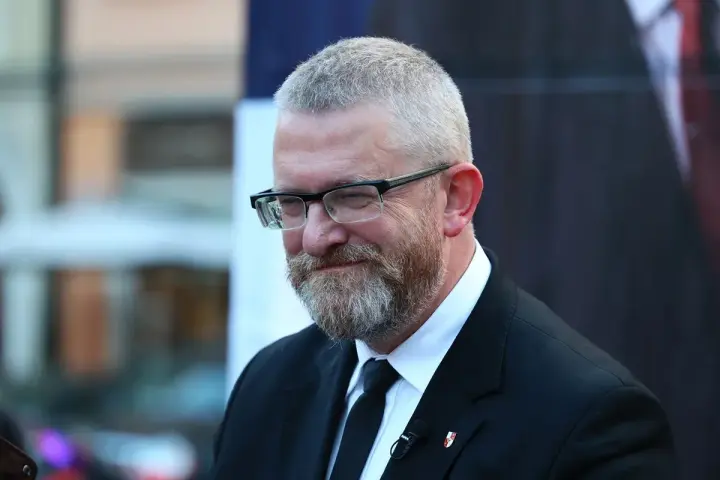 Grzegorz Braun, w okularach i z brodą, w czarnym garniturze, z mikrofonem przypiętym do klapy.