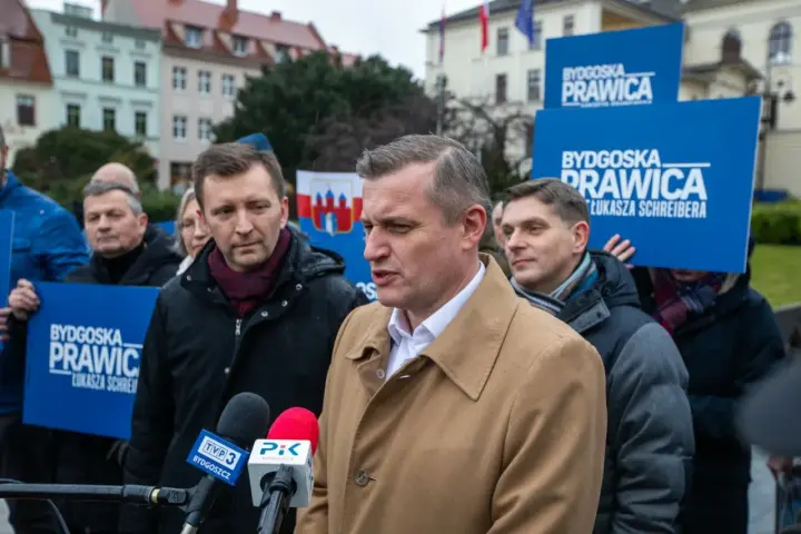 Mężczyzna w płaszczu przemawia do mikrofonów. W tle ludzie z banerami "Bydgoska Prawica". Chcesz wiedzieć, jak zostać politykiem Konfederacji?