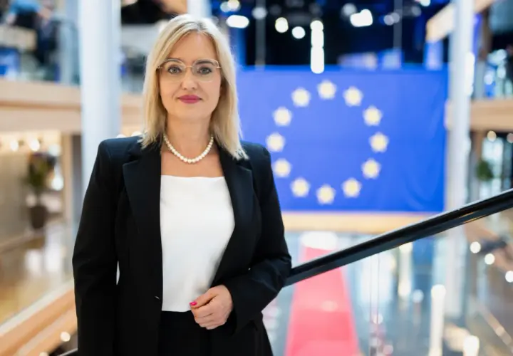 Magda Adamowicz, w eleganckim stroju, stoi przed flagą Unii Europejskiej. Jej wiek nie jest widoczny, ale emanuje pewnością siebie.