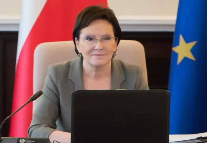 Ewa Kopacz, premier, siedzi przy stole z laptopem, obok flagi Polski i Unii Europejskiej.