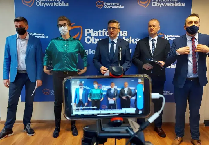 Pięciu mężczyzn w garniturach stoi przed niebieskim banerem z logo Platformy Obywatelskiej. Na pierwszym planie widać telefon nagrywający wydarzenie.