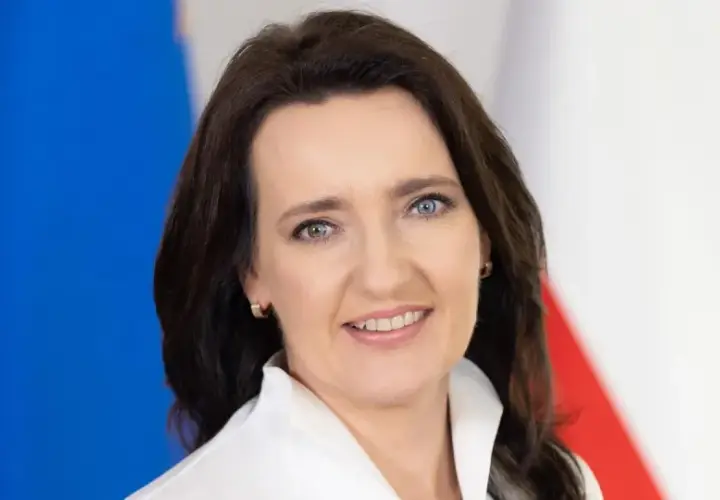 Marzena Okła-Drałowicz, uśmiechnięta kobieta z ciemnymi włosami, na tle biało-czerwonej flagi.