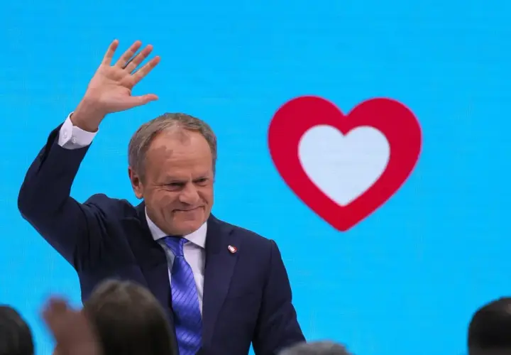 Donald Tusk macha do tłumu na tle niebieskiego ekranu z czerwono-białym sercem, symbolem Koalicji Obywatelskiej.