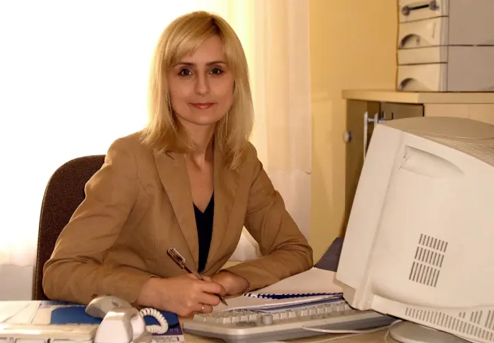 Katarzyna Matusik w lipcu pracuje przy biurku, pisząc coś na papierze. Obok niej komputer i telefon.
