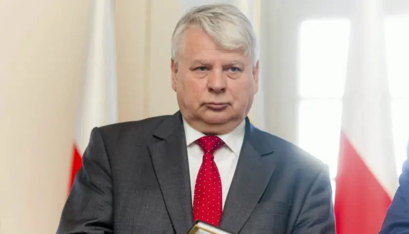 Bogdan Borusewicz w garniturze i czerwonym krawacie, na tle flagi Polski.