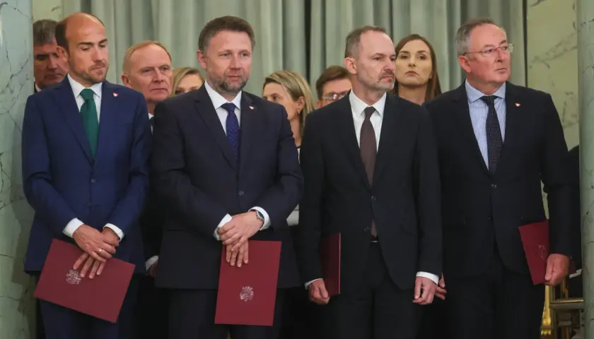 Mężczyźni w garniturach, być może posłowie, stoją w rzędzie, trzymając czerwone dokumenty.