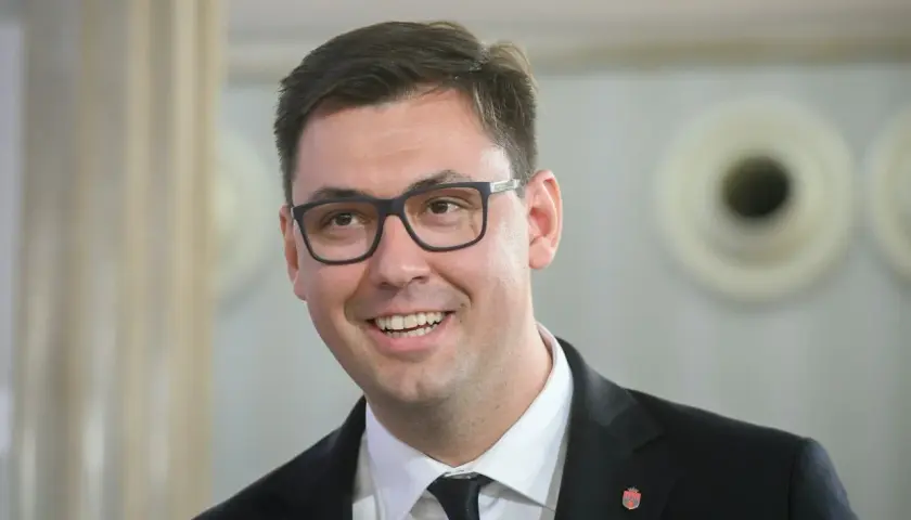 Uśmiechnięty Konrad Frysztak w okularach, w garniturze. Jego wykształcenie pozwala mu na pewność siebie w każdej sytuacji.