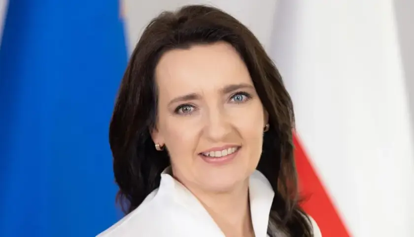 Marzena Okła-Drałowicz, uśmiechnięta kobieta z ciemnymi włosami, na tle biało-czerwonej flagi.