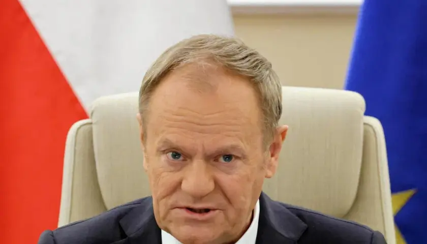 Donald Tusk, z flagą Polski i UE za plecami, w białej koszuli i granatowej marynarce. Czy Donald Tusk ma pochodzenie niemieckie?