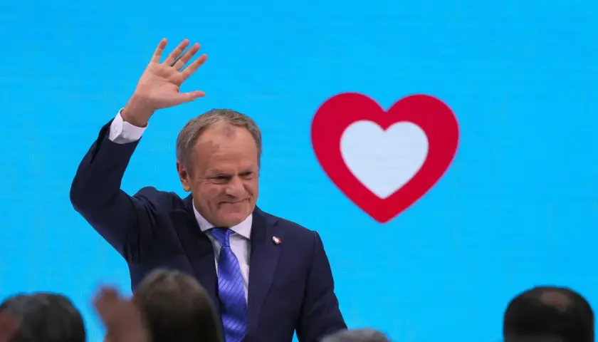 Donald Tusk macha do tłumu na tle niebieskiego ekranu z czerwono-białym sercem, symbolem Koalicji Obywatelskiej.