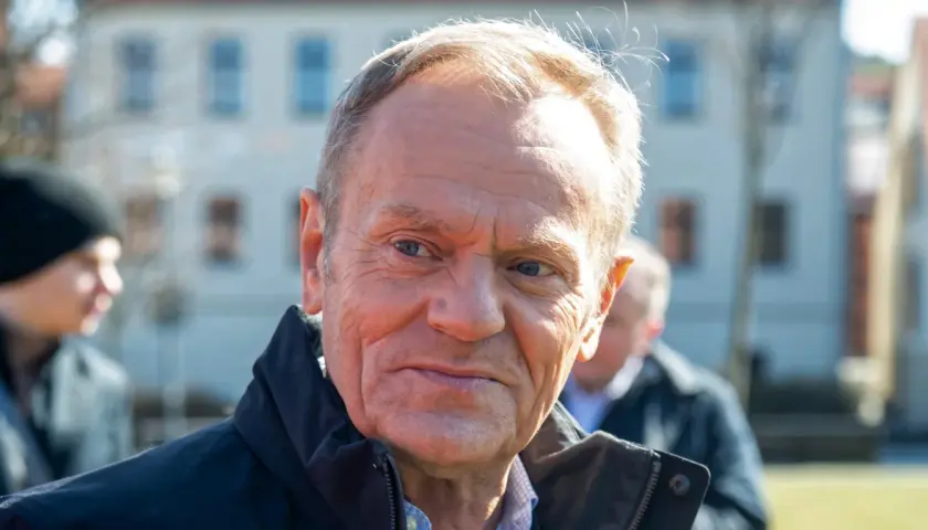 Portret Donalda Tuska w czarnej kurtce, z innymi osobami w tle.