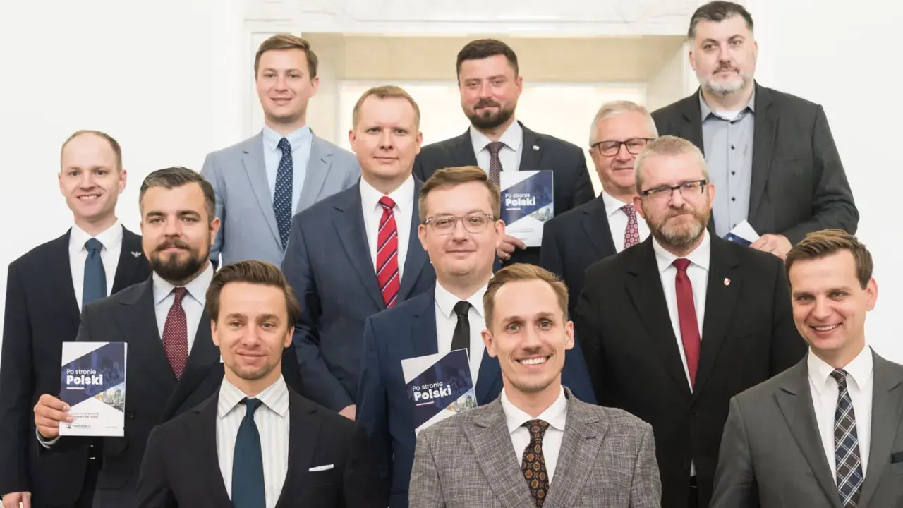 Grupa mężczyzn w garniturach, jeden trzyma broszurę "Po stronie Polski". To członkowie Konfederacji, partii politycznej.