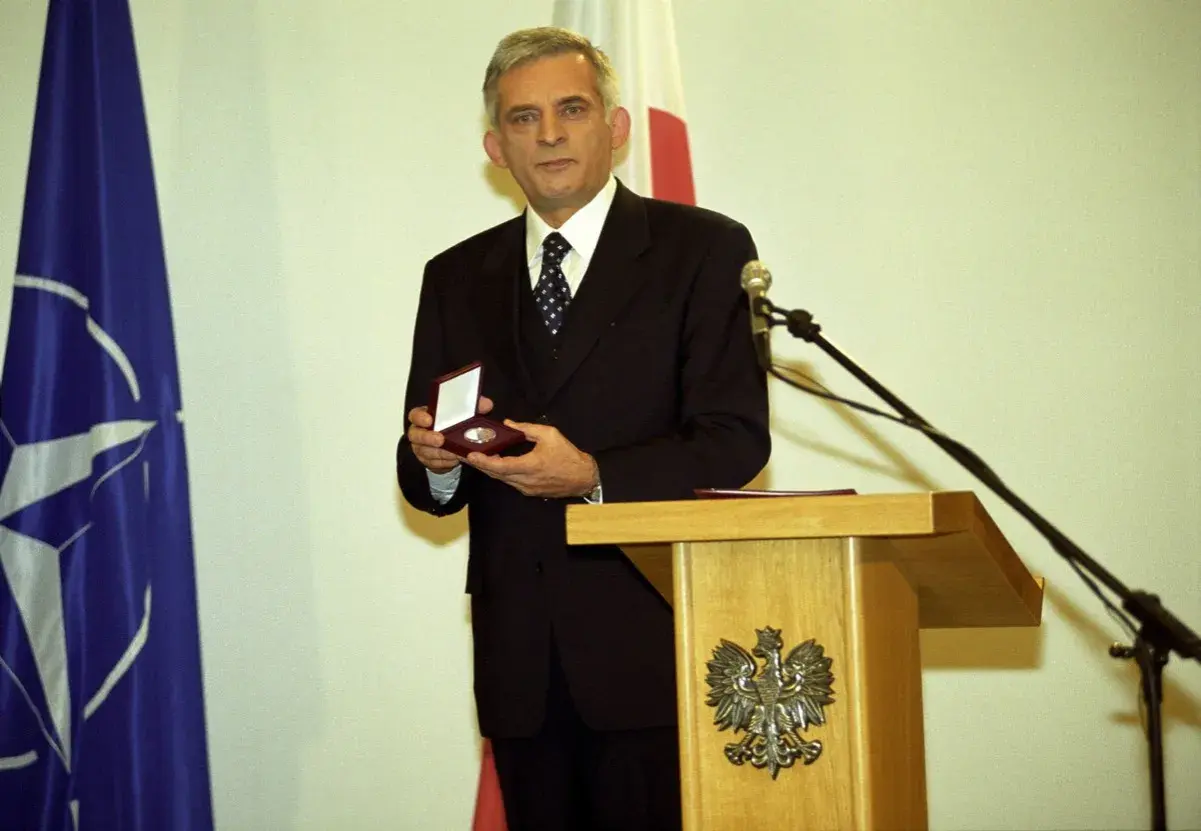Jerzy Buzek, premier Polski, prezentuje medal. Obok flagi NATO i Polski.