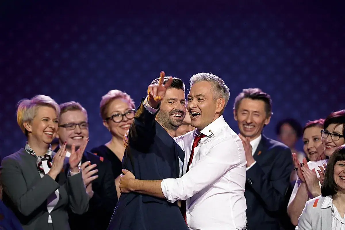 Dwóch mężczyzn, jeden z nich to partner Biedronia, ściska się na scenie, podczas gdy inni ludzie klaszczą.