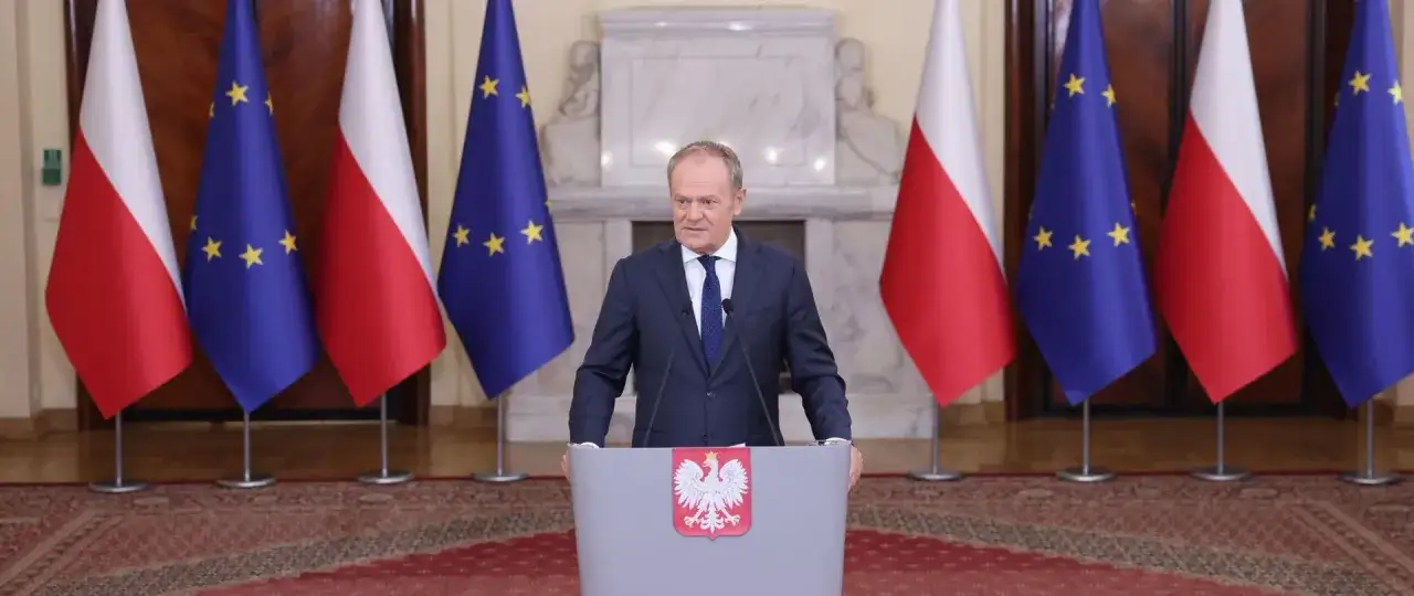 Donald Tusk przemawia na tle flag Polski i Unii Europejskiej.