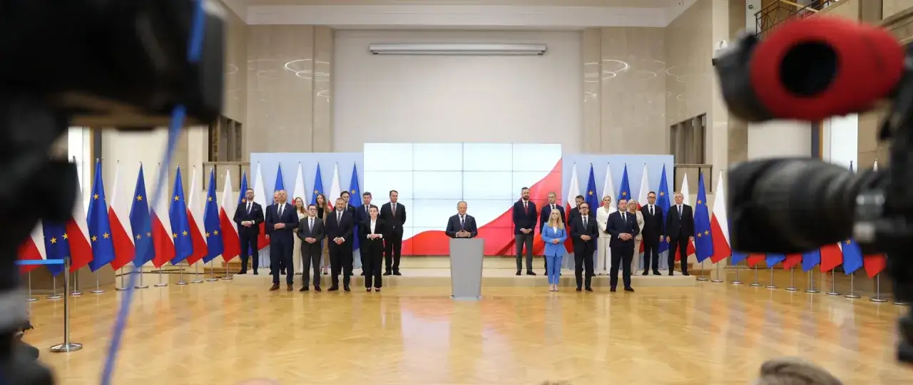 Grupa osób w garniturach i sukienkach stoi przed flagami Polski i UE. W tle ekran z biało-czerwoną flagą. Kto jest premierem Polski?