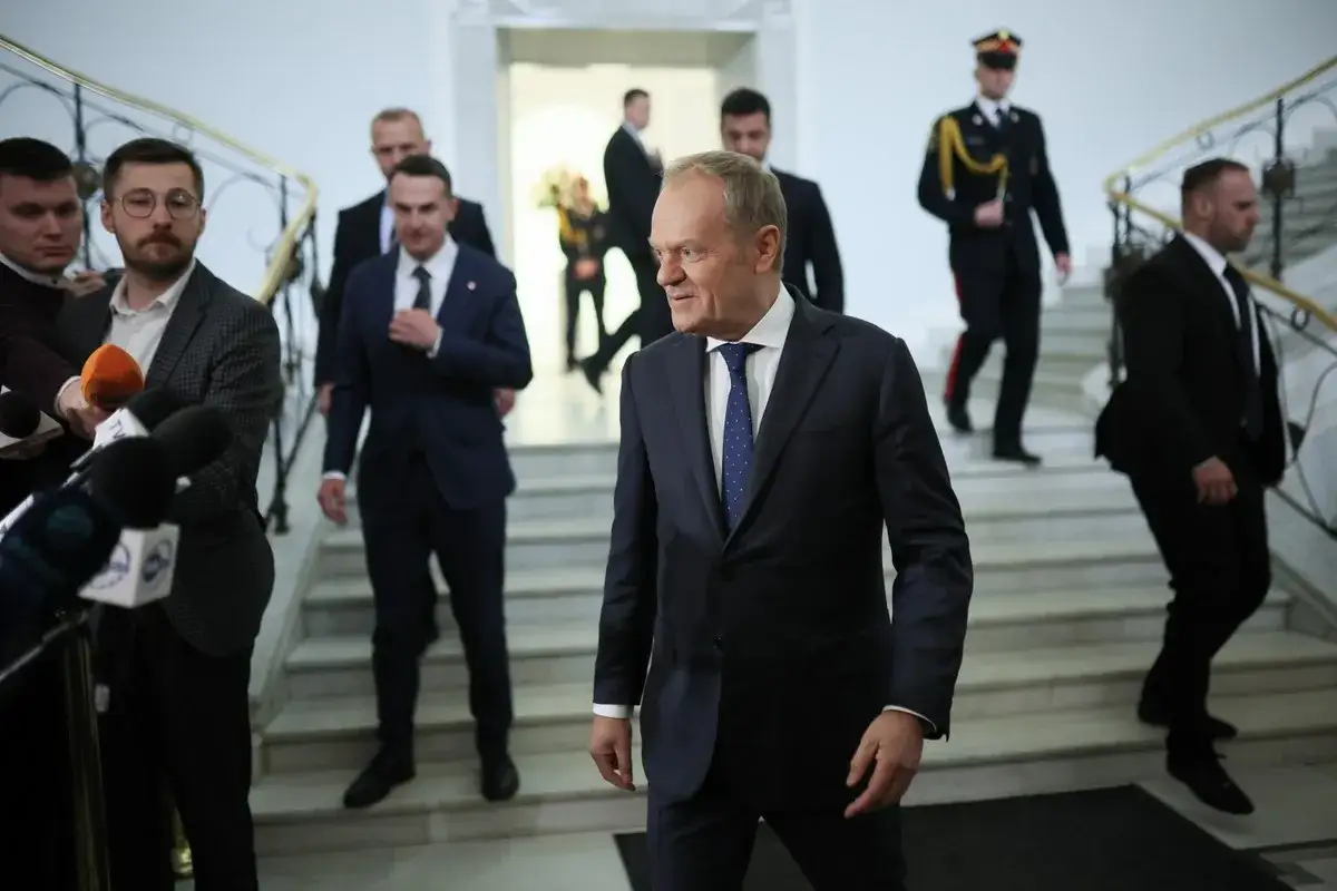Donald Tusk w garniturze, otoczony przez ochronę i dziennikarzy, idzie po schodach. Widać poparcie dla Tuska wśród zgromadzonych.
