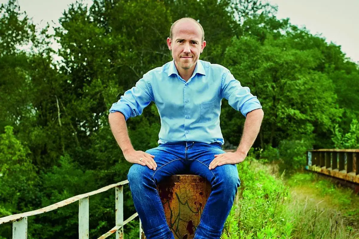 Borys Budka, w wieku, w którym widać doświadczenie, siedzi na tle zieleni.