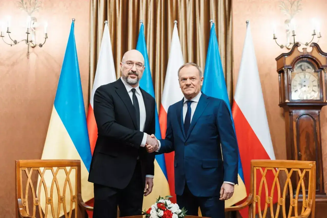 Donald Tusk wita premiera Ukrainy w Brukseli. Na tle flag Polski i Ukrainy politycy ściskają sobie dłonie.