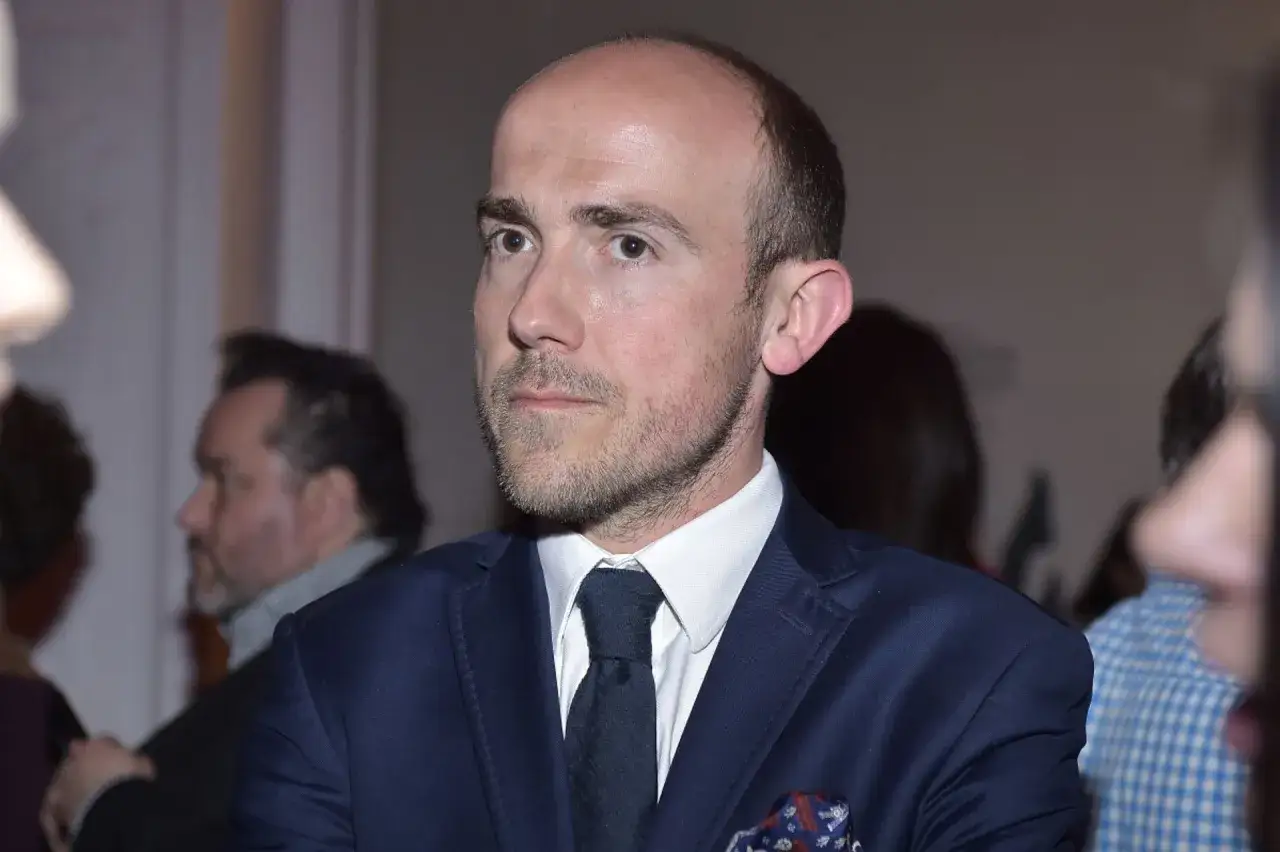 Borys Budka, w eleganckim granatowym garniturze i z charakterystyczną brodą, w otoczeniu innych osób. Jego wiek jest trudny do określenia na podstawie samego zdjęcia.