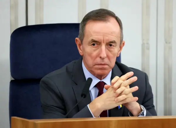 Tomasz Grodzki w garniturze, z rękami złożonymi na piersi, siedzi przy stole. Na klapie marynarki przypinka Poczty Polskiej.
